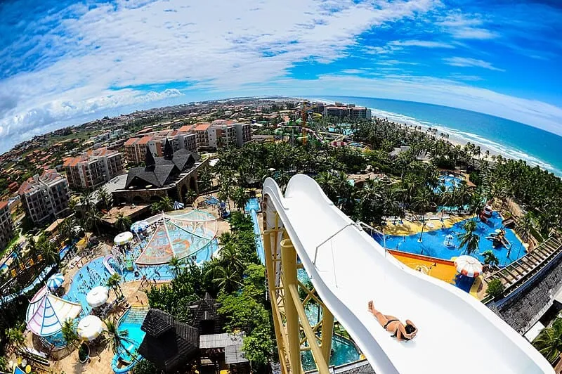 Foto do Parque Aquático Beach Park, em Fortaleza, Ceará. É possível ver um enorme tobogã com uma mulher escorregando pela água. O céu está bem azul e ao fundo é possível ver todo o parque com suas piscinas azuis e vegetação super verde. Ao longe, podemos ver a praia.
