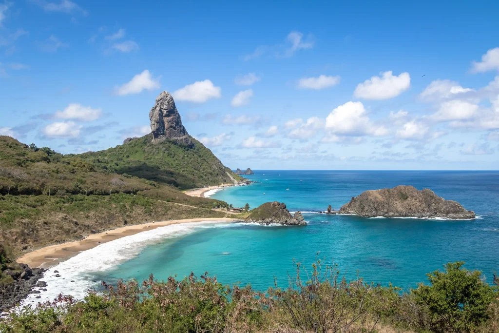 Foto da praia de Fernando de Noronha com as águas bem azuis e cristalinas. O céu está azul. A pedra famosa ao fundo e vegetação verde ao redor.
