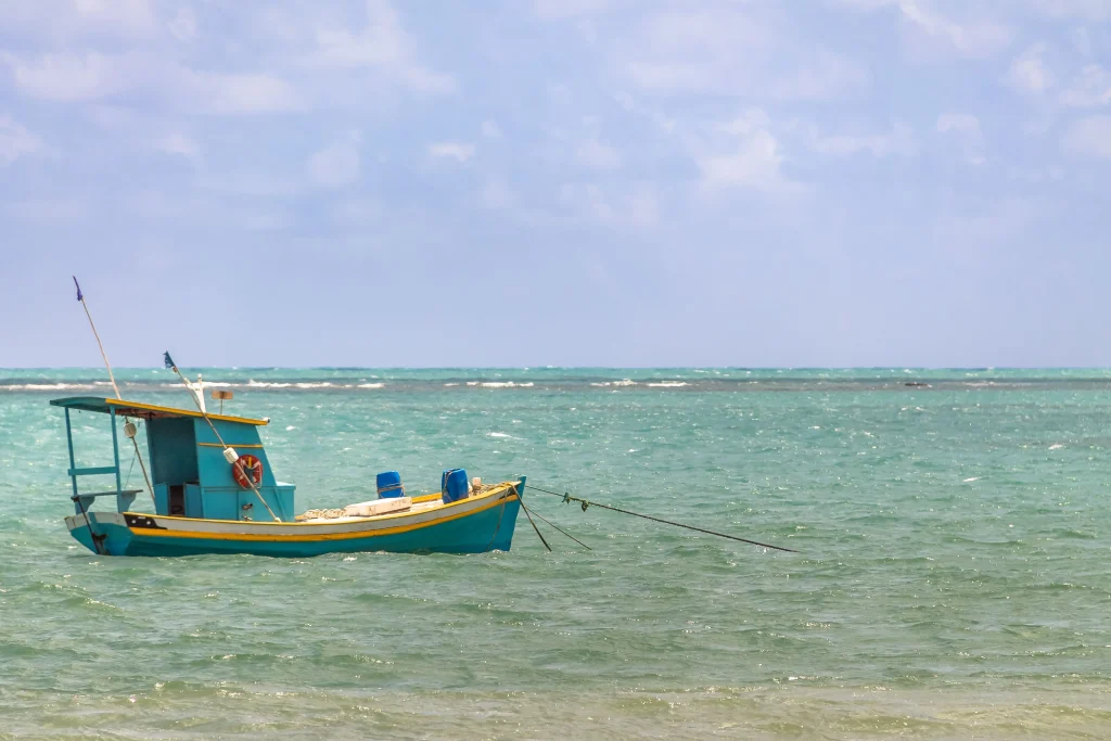Foto de uma das praias do Rio Grande do Norte. As águas são bem azuis e o céu também está azul. No meio do mar encontra-se um barco azul e amarelo estacionado.