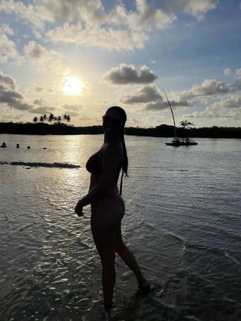 Foto de uma mulher ao centro da foto com os pés no mar. O céu está azul e o sol forte. Há sombra sobre a mulher. Ela sorri e está de óculos de sol.