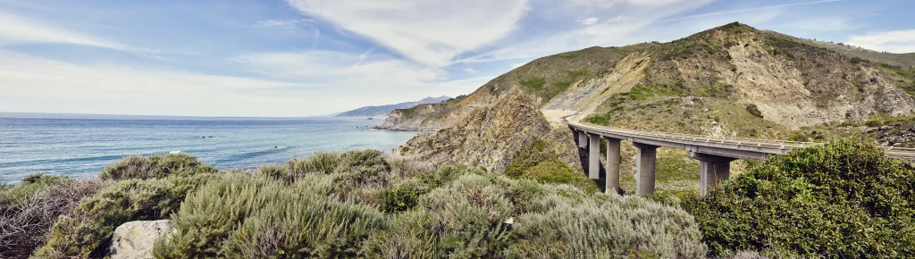 Foto de uma ponte da Big Sur, na Califórnia, Estados Unidos. Ao fundo tem nuvens acima do mar, o mar com ondas, uma faixa de areia, o céu bem azul e a vegetação cobrindo toda a montanha.