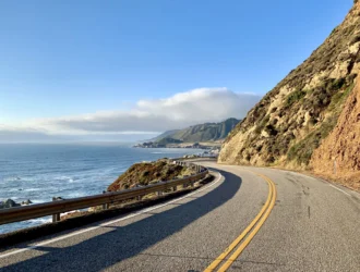 Foto de uma ponte da Big Sur, na Califórnia, Estados Unidos. Ao fundo tem nuvens acima do mar, o mar com ondas, uma faixa de areia, o céu bem azul e a vegetação cobrindo toda a montanha.