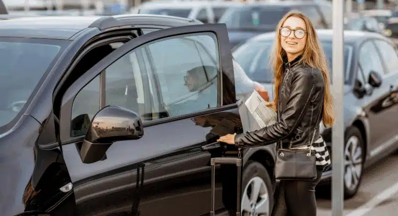 mulher loira com óculos abrindo a porta de um carro preto enquanto segura uma mala
