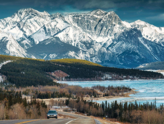 Carro vindo em uma estrada das montanhas rochosas do Canadá com neve no topo e um lago do lado